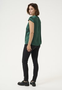 Satin green top with short sleeves, rounded back hem, and a keyhole detail at back, paired with black fitted pants and chunky black shoes.