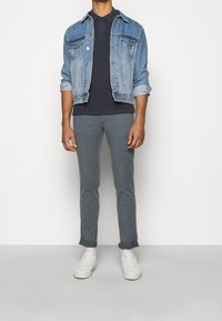 Veste en denim sur polo marine, pantalon gris slim-fit et baskets blanches. La veste a des poches poitrine boutonnées et des manches retroussées.