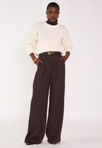 Maglione a maglia testurizzata color crema abbinato a pantaloni marrone a vita alta e gamba larga, con una cintura marrone e una fibbia dorata, braccialetti lucidi.