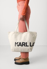 Osoba w koralowych spodniach i brązowych botkach trzyma dużą beżową torbę z czarnym napisem "KARL LAG" na białym tle.