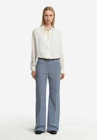 Vit långärmad blus med knappar och krage, tillsammans med ljusblå vida byxor, kompletterad med ett svart bälte och skor.