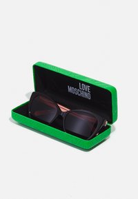 Juodų per didelių saulės akinių žaliai įspaustame dėkle su "LOVE MOSCHINO" ženklu. Lęšiai tamsūs, o rėmelis turi geometrinę formą.