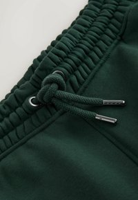 Tissu vert foncé avec une taille élastique et un cordon noué, doté d'embouts et d'œillets métalliques.