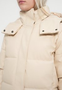 Casaco acolchoado bege com gola alta, detalhes de botões de pressão nos ombros e textura acolchoada. Acabamento suave e macio.