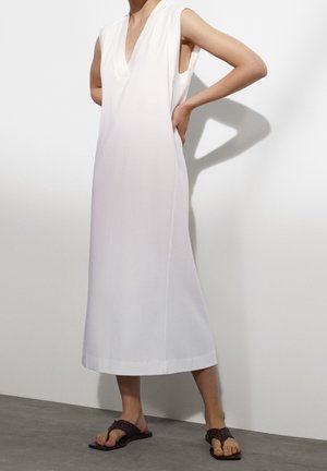 Robe blanche sans manches avec un décolleté en V, silhouette droite et longueur mi-mollet. Portée avec des sandales sombres et texturées. Tissu lisse et design simple.
