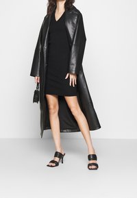 Manteau long en cuir noir sur une robe tricotée ajustée de couleur sombre. Sandales à talons ouverts noires. Texture lisse avec une silhouette épurée. Accessoires non détaillés.