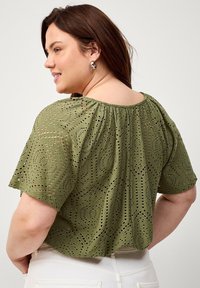 Oliven grøn bluse med korte ærmer, åbent arbejde detaljer, elastisk hals og en afslappet pasform. Stoffet har cirkulære mønstre og en blød tekstur.