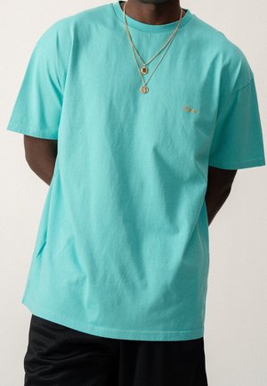 T-shirt en coton turquoise avec un col rond et des manches courtes, présentant un petit logo brodé en or sur la poitrine.