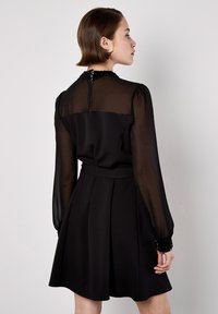 Vestido negro con parte superior de manga larga transparente y falda lisa, con plisados y puños adornados. Cierre con cremallera en la parte posterior, silueta refinada.