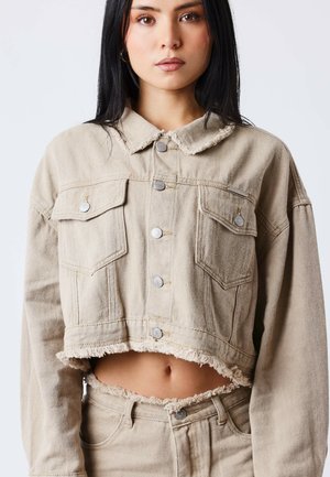 Femme portant une veste en jean beige courte aux bords effilochés et un pantalon taille haute assorti, debout devant un fond clair uni.