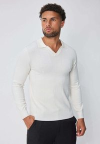 Pull blanc à manches longues avec col en V, présentant une texture côtelée verticale et une coupe décontractée, associé à un pantalon foncé.