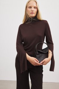 Braunes Strickoberteil mit ausgestellten Ärmeln, kombiniert mit einer schwarzen Handtasche mit rundem Griff und weicher Textur. Das Model hält die Tasche vor sich.