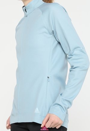 Woman wearing light blue long-sleeve zip-up sports jacket with Adidas logo near hem and black pants with pink Adidas logo.