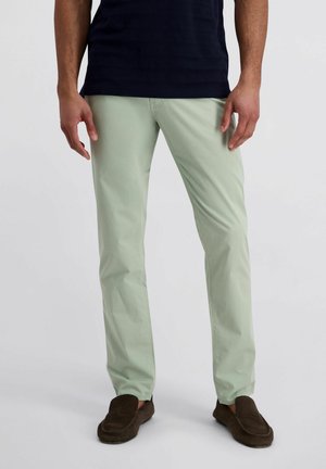 Mann trägt hellgrüne gerade geschnittene Hosen, dunkelbraune Loafers und ein schwarzes Hemd, steht vor einem schlichten hellen Hintergrund.