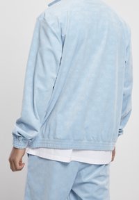 Světle modrá tepláková bunda s elastickými manžetami a pasem, zdobená jemným tónovaným vzorem oblouků. Měkká textura, klasický design.