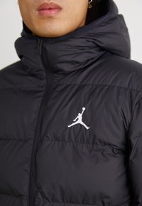 Jachetă neagră puffer cu guler înalt, dotată cu panouri cusute și un logo alb Jordan pe pieptul stâng. Textura este netedă și ușor strălucitoare.