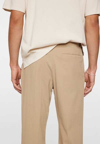 Beige strukturierte Hose mit einer Gesäßtasche, kombiniert mit einem hellbeigen Kurzarmhemd. Der Stoff hat ein dezentes gewebtes Muster.