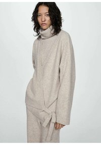 Béžový pletený sveter s vysokým rolákom a prekrývajúcim dizajnom, s bočným viazaním. Mäkkučká textúra a široké rukávy.