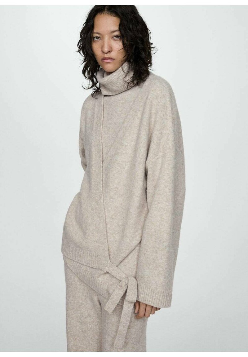 Béžový pletený sveter s vysokým rolákom a prekrývajúcim dizajnom, s bočným viazaním. Mäkkučká textúra a široké rukávy.