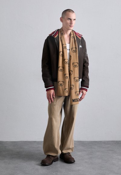 Chaqueta marrón con acentos a rayas rojas y blancas, camiseta de color crema, bufanda color beige con patrones de oso, pantalones beige y botas marrones. Fondo sencillo.
