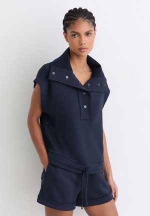 Kvinde med flettet hår iført en navy ærmeløs top med knapper og matchende shorts med snøre, stående mod en ensfarvet hvid baggrund.