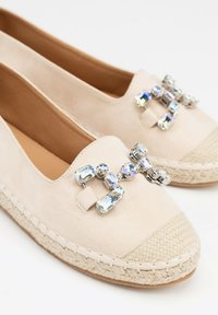 Espadrillas beige in suede con suola di juta intrecciata, caratterizzate da un accento decorativo in strass frontale. Texture liscia e forma arrotondata.