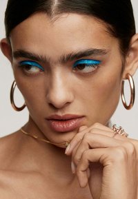 Primer plano de un rostro con delineador de ojos azul brillante, pendientes de aro dorados y anillos en capas. La piel es suave, los labios son de un tono nude y el cabello es lacio.