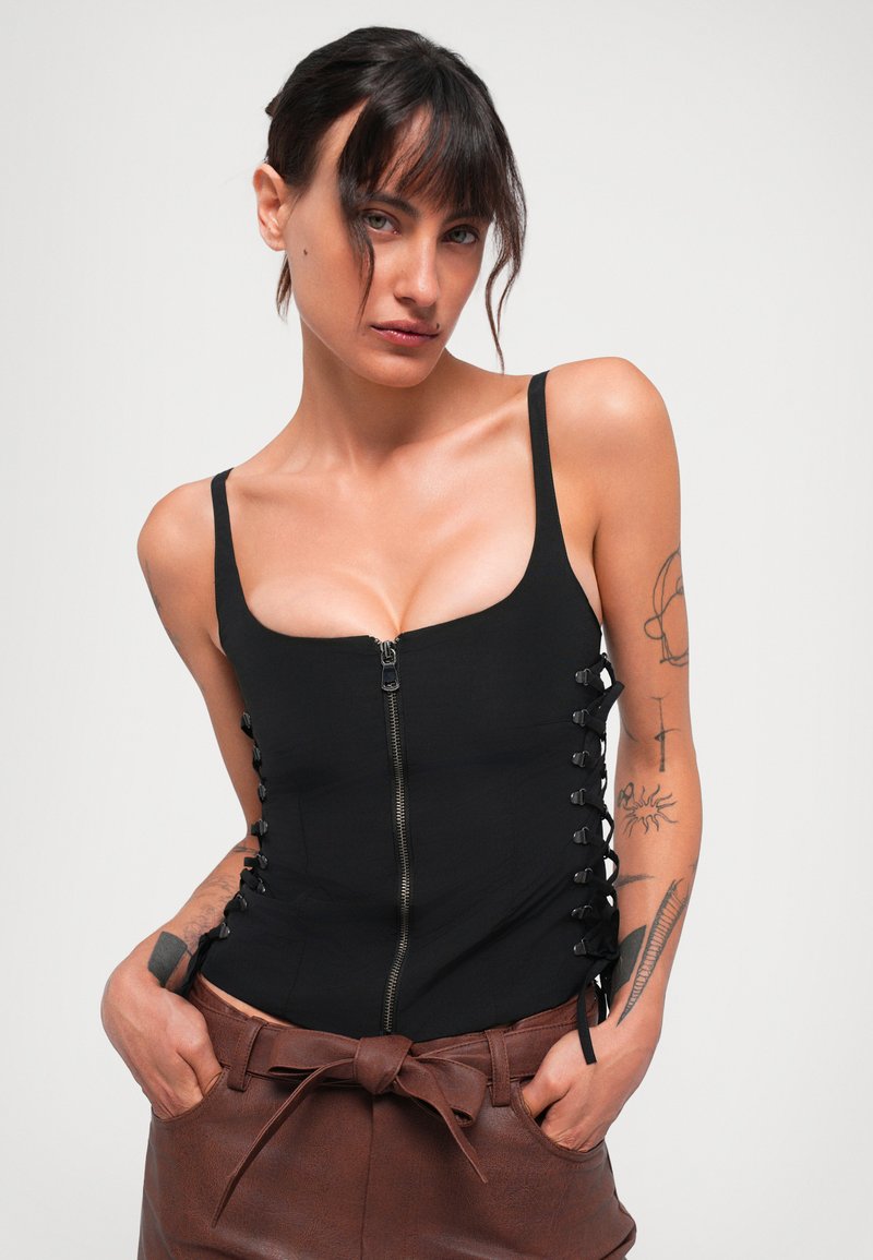 Zwarte fitted bodysuit met een ritssluiting aan de voorkant, accenten van zijveters, brede bandjes en een gladde textuur, gecombineerd met hoog getailleerde, bruine leren shorts.