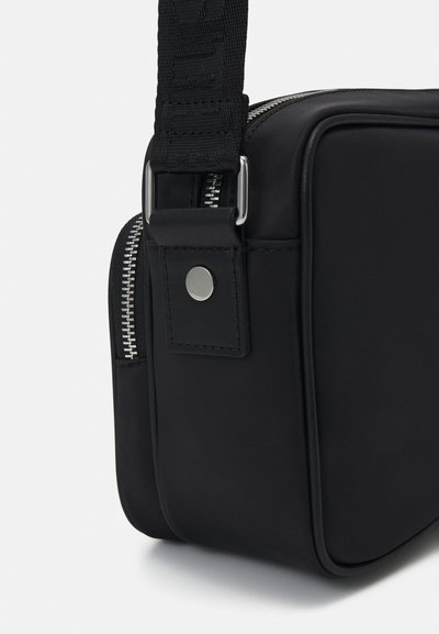 Bolso rectangular negro hecho de un material suave, con detalle de cremallera plateada, cierre de botón a presión y una correa de hombro ajustable texturizada.