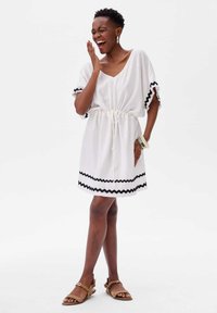 Witte jurk met een V-hals, getailleerde taille en korte mouwen met zwarte ruches. G gedragen met bruine sandalen en meerdere armbanden.