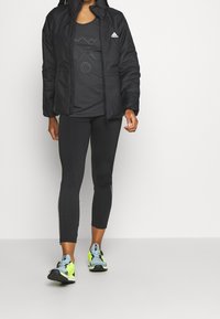 Giacca imbottita nera con cappuccio, canotta grafica grigia scura, leggings neri con strisce laterali e scarpe sportive blu-gialle.