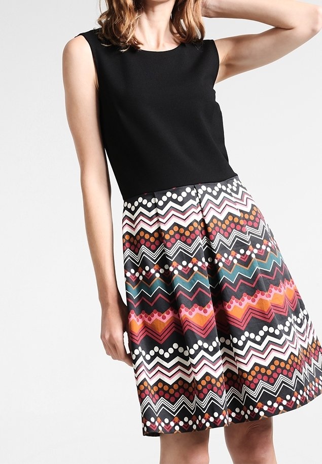 Femme portant une robe noire sans manches avec une jupe à motif zigzag et pois colorés dans des tons rose, orange, sarcelle et blanc.