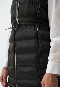 Schwarze Steppweste mit horizontalem Quiltmuster, silbernem Reißverschluss und verstellbarem Kordelzug in der Taille. Kombiniert mit einem weißen Langarmshirt.