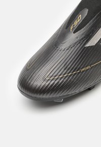 Bota de fútbol negra con textura acanalada, que presenta acentos dorados y un diseño alargado. Logo "F50" y "SPRINTGRID" visibles en la parte superior.