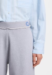 Άτομο που φοράει γαλάζιο πουκάμισο με ρίγες και γκρι παντελόνι φόρμας με φθαρμένη μέση και ένα μικρό μπλε κεντημένο λογότυπο με χαμογελαστό προσωπάκι.