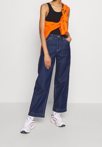 Persona che indossa dei jeans a gamba larga blu scuro, una canottiera nera, una felpa arancione annodata intorno alle spalle e sneakers bianche, in piedi contro uno sfondo semplice.
