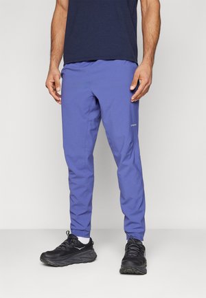 Mand iført blå træningsbukser, hvide sokker, sorte løbesko og en mørk navy t-shirt står foran en ensfarvet baggrund.