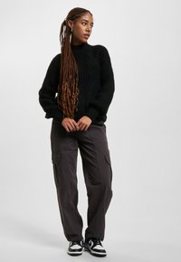 Fülliger schwarzer Strickpullover mit hohem Kragen und langen Ärmeln, kombiniert mit dunkelgrauen Cargo-Hosen mit Seitentaschen und schwarzen Sneakers.