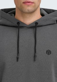 Felpa grigia realizzata in tessuto morbido, con cappuccio regolabile tramite un laccio, ganci neri e un piccolo logo a cubo nero sul petto.