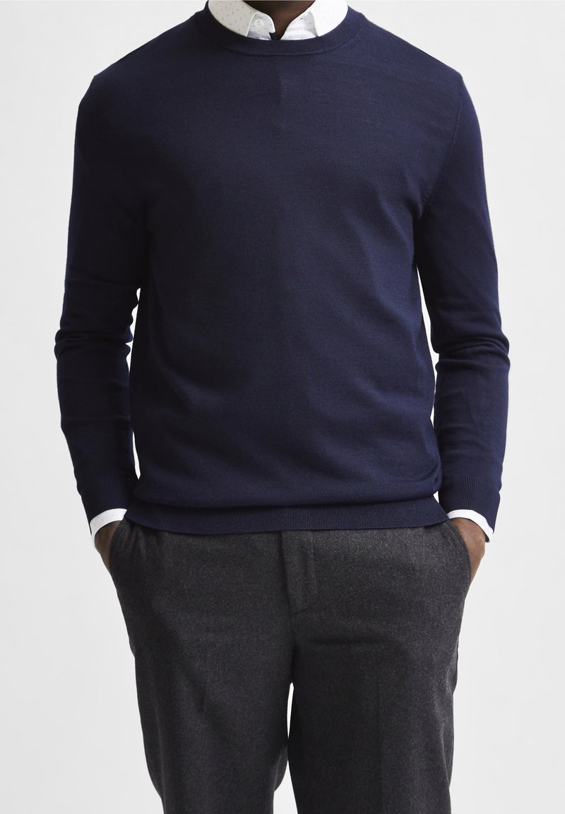 Mörkblå stickad tröja med rund halsringning, ribbade ärmslut och nederkant. Bärs över en vit skjorta med prickar, kombinerad medgrå byxor.