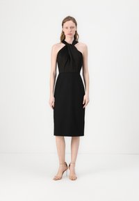 Crna halter haljina s uvijenim detaljem na tijelu, uskim strukom i do koljena dugom donjim dijelom. Model nosi puderaste sandale s remenom oko gležnja.