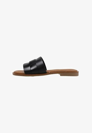 Sandalia plana negra con una correa superior ancha y textura suave. Suela marrón claro con un diseño minimalista, sin relieve y sin herrajes visibles.