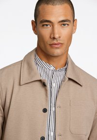 Beige Baumwolljacke mit Kragen, schwarzen Knöpfen und einer Brusttasche, über einem weiß und grau gestreiften Hemd getragen.