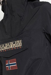 Schwarze Nylonjacke mit hohem Kragen, Reißverschluss und einem großen Logo-Patch mit gestickter norwegischer Flagge am Ärmel.