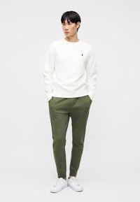 Νέος άνδρας που στέκεται με τα χέρια στις τσέπες, φορώντας λευκό πουκάμισο με μακριά μανίκια, χακί φόρμα και λευκά αθλητικά παπούτσια μπροστά σε απλό φόντο.