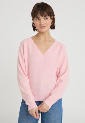 Giovane donna con capelli corti castani che indossa un maglione rosa chiaro a scollo a V e jeans blu, in piedi davanti a uno sfondo bianco semplice.