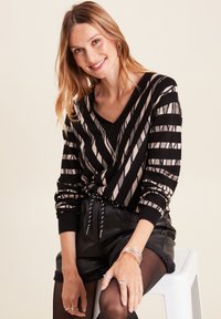 Schwarzer und kremfarbener gestreifter Strickpullover mit V-Ausschnitt, präsentiert diagonalen Mustern und gerippten Bündchen, kombiniert mit schwarzen Leder-Shorts.