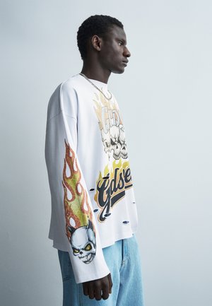 Νεαρός άνδρας με λευκό μακρυμάνικο πουκάμισο με γραφικά φλεγόμενου κρανίου και ανοιχτό μπλε τζιν, στέκεται πλευρικά σε απλό ανοιχτό φόντο.