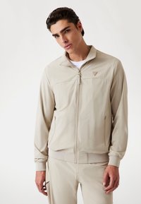 Beige zip-up jakke i et letvægtsmateriale med krave, sidelommer og ribbede manchetter. Har en subtil logo-detalje på brystet.