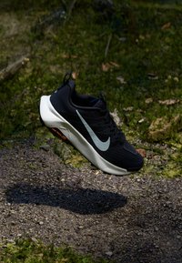 Black athletic shoe with a textured upper, light gray swoosh, and white sole. Features a padded collar and reflective accents.